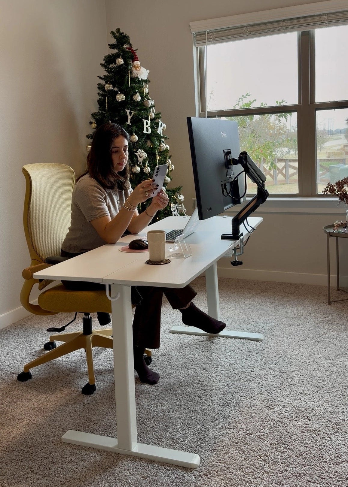 A woman uses her phone at a white Sunaofe standing desk and yellow ergonomic chair in a festive home office.