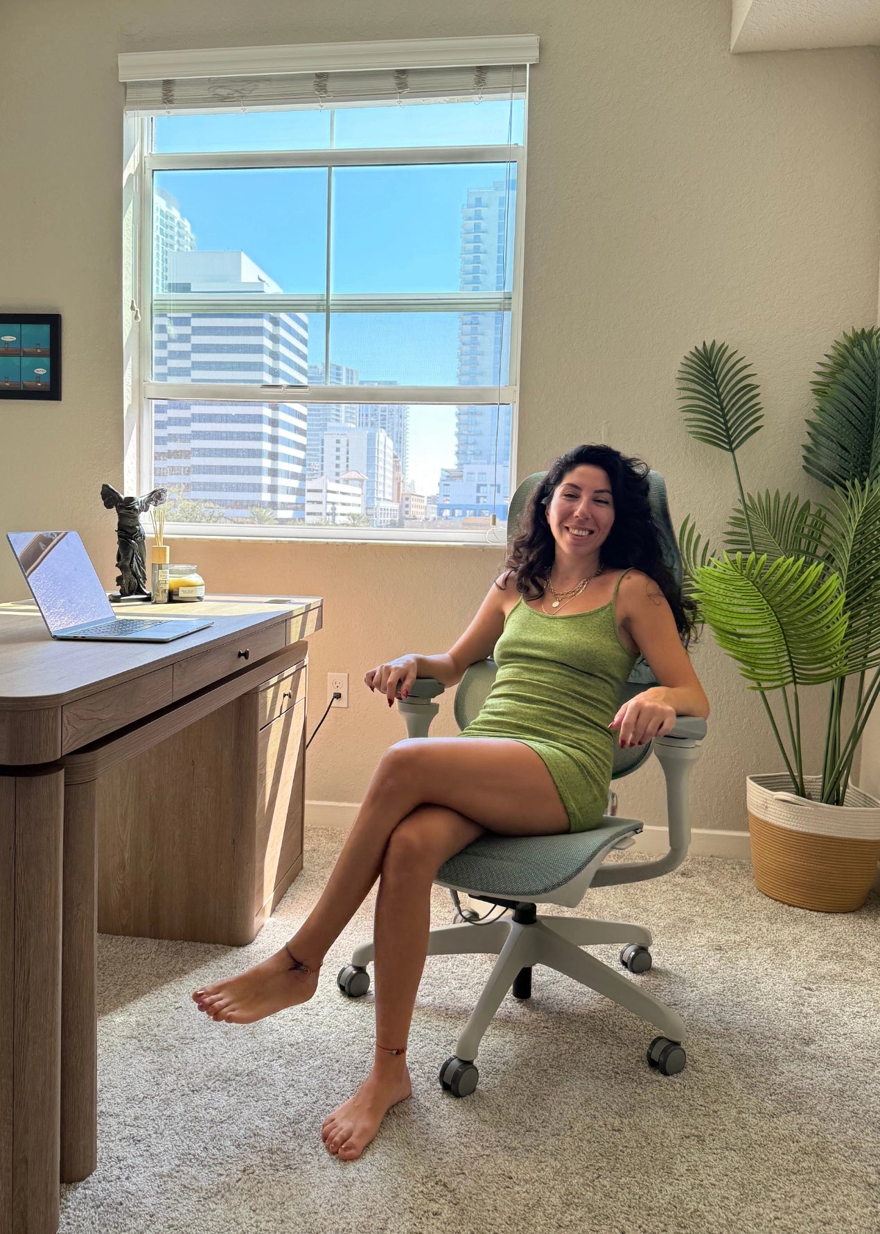 A smiling woman sits comfortably in a Sunaofe ergonomic office chair in a bright, sunlit home office.